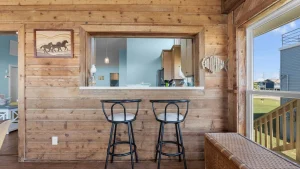 Rustic wood-wall kitchen pass-through with a white counter, two black metal bar stools, and a wicker bench; coastal decor and blue kitchen visible beyond.