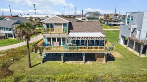 Aerial view of a coastal two-story home with a large wooden deck and turquoise chairs on the balcony and yard.