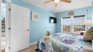 Bedroom with light blue walls, queen bed, ocean-themed quilt, and folded towels on bed; stairs visible through open door left.
