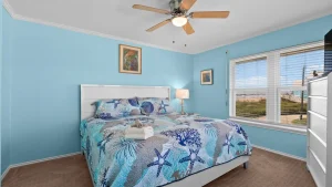 Coastal-themed bedroom with light blue walls, a white bed frame and sea-themed quilt, and a large window showing a beach view at right.