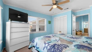 Bright blue bedroom with a white dresser supporting a flat-screen TV and decorative seagull figurine, next to a window with blinds and a patterned ocean-themed bedspread.