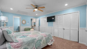 Coastal-style bedroom with a ceiling fan, light blue walls, and white wainscoting; a wall-mounted TV sits above white louvered closet doors.