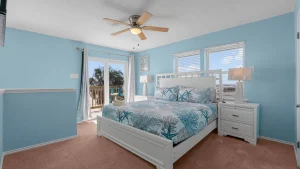 Coastal bedroom with light blue walls, white bed frame, and ocean-themed bedding facing a balcony door. Elements include two white nightstands and a ceiling fan with wooden blades.