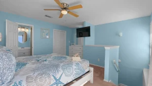Coastal bedroom with light blue walls, white furniture, and a ceiling fan; bed in foreground with sea-pattern quilt and a wall-mounted TV.
