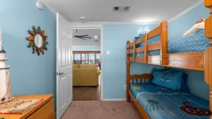 Small blue bunk room with wooden twin bunks, blue bedding, and a doorway to a living area seen in the background.
