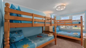 Two wooden bunk beds in a bright blue kids' room with ocean-themed quilts and pillows, near a window with blinds