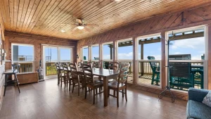 Sunroom with a wood-paneled ceiling and walls, a long wooden dining table, and chairs, plus large windows looking out to a deck and blue sky/ocean.