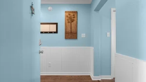 Bright blue hallway with white beadboard wainscoting, a palm tree wall art piece, a small mirror shelf, and coat hooks on the wall.