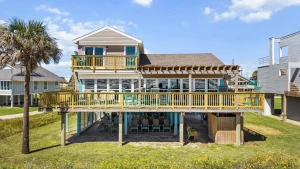 Two-story beach house with a large wooden deck and turquoise chairs, under a bright blue sky with a palm tree nearby.