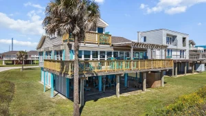 Two-story beach house with wraparound wooden deck, teal accents, and a tall palm tree on a sunny day in a coastal neighborhood.