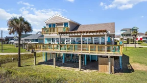 Beach house with wraparound decks and turquoise chairs, raised on stilts, under a blue sky with a palm tree nearby.