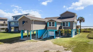 Coastal house on stilts with blue accents, facing the ocean and a palm tree to the right; outdoor seating under the deck.