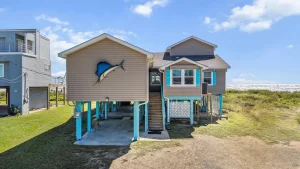 Beached elevated house with a blue marlin wall sculpture, beige siding, turquoise supports, and stairs to the entrance near the ocean.