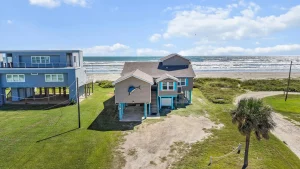 Beachfront houses by the ocean: blue multi-story home on the left and a beige house with blue trim near the shore; palm tree in foreground and blue sky with clouds.