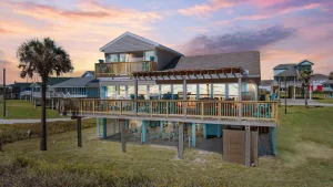 Two-story beach house on stilts with a wraparound deck and turquoise chairs at sunset.