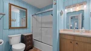 Blue-walled bathroom with a glass shower enclosure, wooden vanity, and a framed mirror above the toilet as part of a coastal-style bath suite.