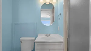Small blue bathroom with a white sink, toilet, and oval mirror reflecting a window and sky outside.
