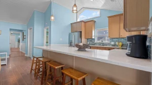 Bright kitchen with blue walls, white countertop island, wooden stools, and stainless fridge under pendant lights.