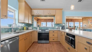 Coastal kitchen with light wood cabinets, teal tile backsplash, and stainless appliances; open doorway to a wood-paneled living area with an ocean view.