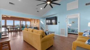 Coastal living room with blue walls, mustard sofas, wood floors, and a ceiling fan; TV mounted on the wall near a doorway to a bedroom.