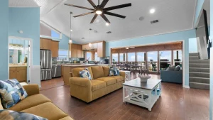 Open-plan living room and kitchen in a coastal home with blue walls, white ceiling, and a large deck visible through glass doors.