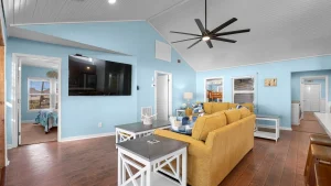 Bright living room with light blue walls, a mustard sofa, white coffee tables, and a wall-mounted TV. A ceiling fan hangs from the white beamed ceiling above.