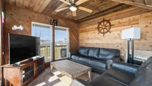 Cozy cabin living room with wood paneling, black leather sofa set, wooden coffee table, and a mounted TV on a wooden cabinet. Neon Howdy sign by window.