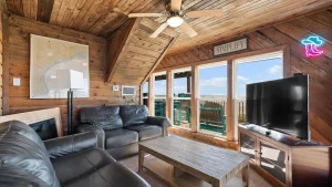 Rustic, wood-paneled living room with black leather seating, a coffee table, and large windows overlooking a beach deck.