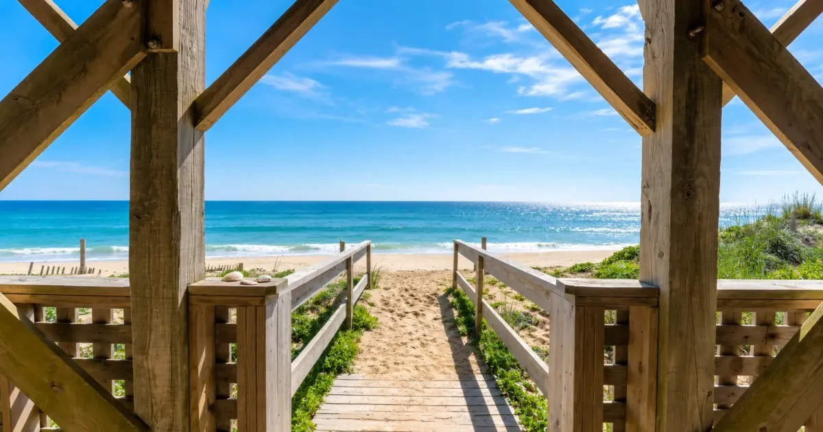 View from a wooden gazebo toward a sandy beach and calm blue sea under a clear sky.